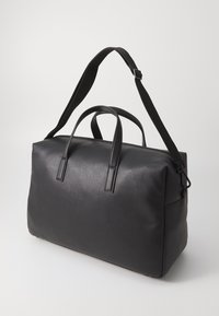 Sac de voyage en cuir noir avec deux poignées supérieures et une bandoulière réglable. Surface texturée avec des accents lisses. Forme rectangulaire.