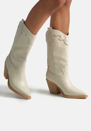 Botas de cuero crema suave con bordes decorativos en forma de escallo, puntera puntiaguda y un tacón de bloque de madera con ranuras verticales.