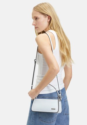 White leather crossbody bag with black trim, triangular shape, adjustable strap, and silver hardware detail. Worn against a denim background.