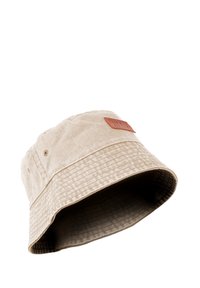 Tan bucket hat made of textured fabric with a wide brim and a leather logo patch on the side. Features stitching patterns along the brim.