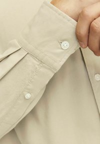 Beige Langarmhemd mit Knopfmanschette, das eine glatte Textur und dezente Nähdetails aufweist. Zwei weiße Knöpfe sind an der Manschette sichtbar.
