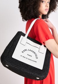 Svart vävd toteväska med vita läderdetaljer. Har logotyp och adress på framsidan, platta handtag, kvadratisk form och rundad botten med hårdvara.