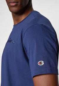 Dunkelblaues Baumwoll-T-Shirt mit kurzen Ärmeln, mit gesticktem Logo auf der Brust und Logo-Patch am Ärmel. Glatte Textur, klassischer Schnitt.
