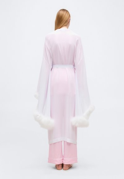 Γυναίκα με μακριά μαλλιά στέκεται ξυπόλητη, φορώντας μια διάφανη λευκή ρόμπα με γούνα στο τελείωμα πάνω από ανοιχτό ροζ παντελόνι πιτζάμας, με την πλάτη προς τα εμάς σε απλό φόντο.