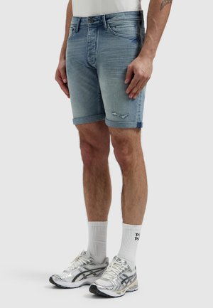 Man met lichtblauwe denim shorts met opgerolde zoom en lichte slijtage, witte sportsokken en witte hardloopschoenen op een effen achtergrond.