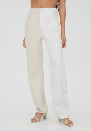 Vrouw draagt high-waist rechte jeans, één pijp beige en de andere wit, gecombineerd met witte sandalen met hak.