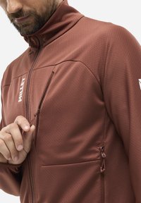 Brown zip-up jacket with textured fabric, featuring a chest pocket. Zipper pulls and a logo on the front are notable design details.