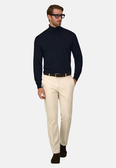 Homme portant un col roulé noir, un pantalon beige, une ceinture noire, des bottes noires et des lunettes, debout avec une main dans la poche.