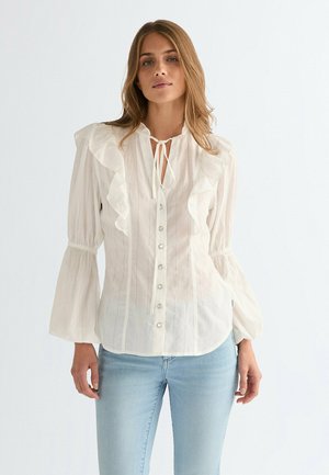 Blusa blanca con detalles plisados, cierre frontal de botones y cuello con cordón. Presenta mangas largas abullonadas y rayas verticales en una tela transparente.