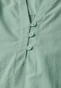 Tessuto di camicia verde menta con scollatura a V, caratterizzato da un patta con tre bottoni coordinati e un delicato dettaglio di cucitura.