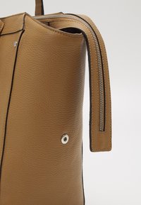 Torba na ramię ze skóry w odcieniu jasnobrązowym o fakturowanej powierzchni, wyposażona w srebrny zamek błyskawiczny, górną rączkę oraz ozdobny zatrzask po boku.