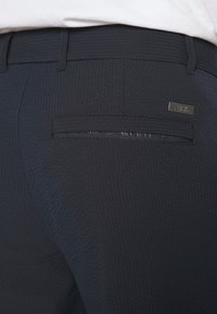Gros plan sur la poche arrière d'un pantalon texturé bleu marine avec une petite étiquette métallique de marque au-dessus de la poche et des passants visibles.