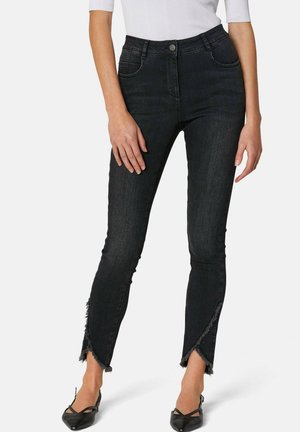 Vrouw draagt zwarte high-waist skinny jeans met asymmetrische rafelige zomen en zwarte platte schoenen met een puntige neus, gecombineerd met een wit topje.