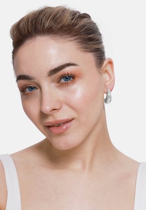 Des boucles d'oreilles en argent en forme de goutte présentées sur un modèle. Les boucles d'oreilles ont une surface lisse et réfléchissante ainsi qu'un design minimaliste.