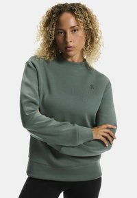 Roheline kapuutsiga särk ümmarguse kaelusega, pikkade varrukatega ja sooniku tekstuuriga. Rinnal väike logo, kombineeritud mustade retuusidega.