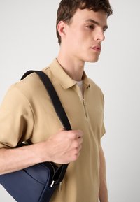 Beige, rövid ujjú pólóing félig zipzárral a gallérnál. Sötét tengerészkék válltáska cipzáras záródással. Simaságot árasztó anyag, minimális dizájn.