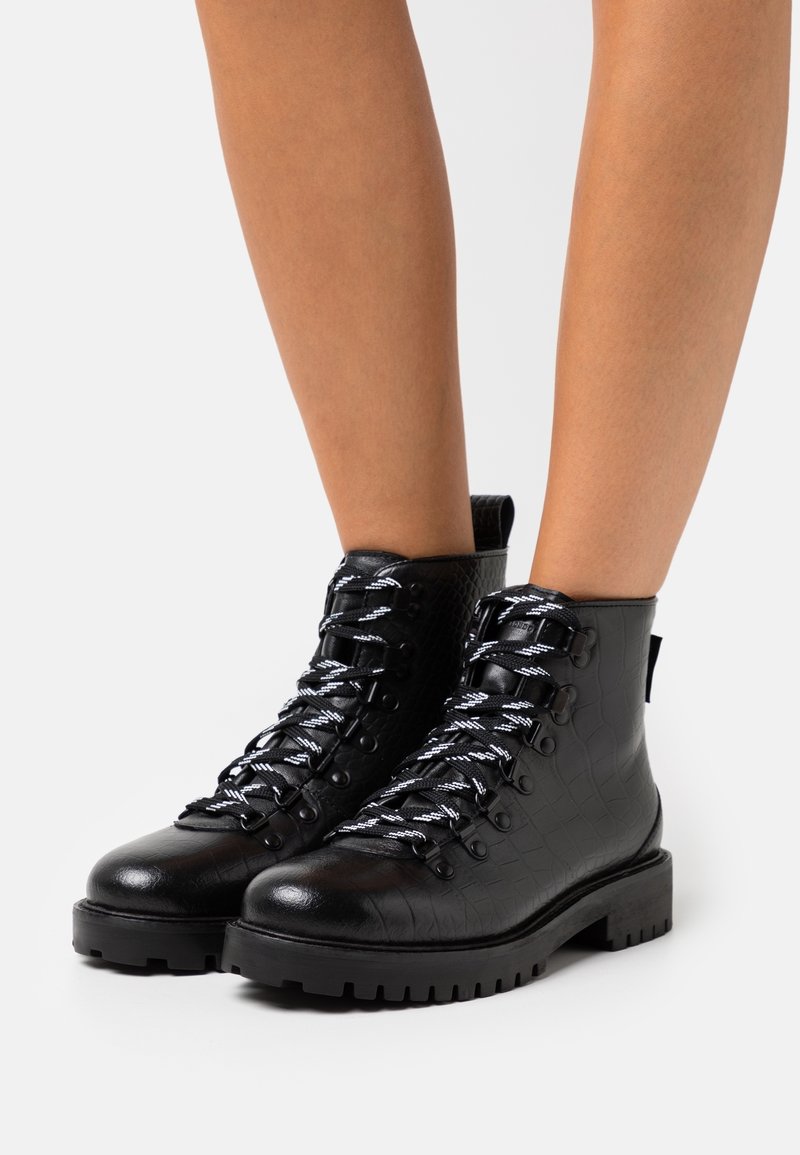 Botas de tobillo de cuero negro con textura de cocodrilo, puntas redondas y cordones en blanco y negro. Cuentan con una suela gruesa y resistente y una lengüeta.