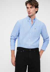 Jasnoniebieska koszula z długim rękawem w cienkie białe i czerwone paski; posiada kieszonkę na piersi oraz małe logo. W połączeniu z czarnymi dżinsami.