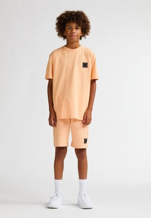 Set van T-shirt en korte broek van katoen in perzikkleur met een zwart logoplaatje. Heeft een ontspannen pasvorm, korte mouwen en opgerolde zomen aan de korte broek.