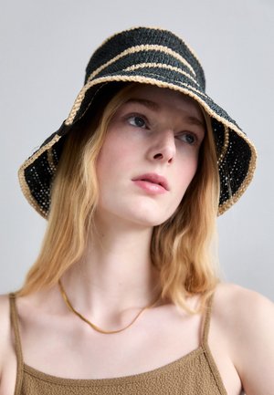 Młoda kobieta o jasnej cerze i blond włosach, nosząca czarno-beżowy pasiasty pleciony kapelusz typu bucket oraz brązowy top na ramiączkach z cienkim naszyjnikiem.