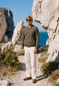 Homme portant des lunettes de soleil, une veste olive, un pantalon blanc et des chaussures marron, debout sur un chemin rocheux avec la mer et des falaises en arrière-plan sous un ciel clair.