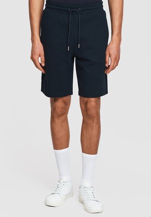 Mężczyzna w czarnych szortach z troczkiem, białych skarpetach sportowych i białych trampkach, stojący na jasnym jednolitym tle.