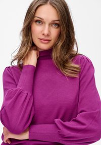 Kobieta z jasnobrązowymi włosami i zielonymi oczami, ubrana w fioletowy sweter z długim rękawem i bufiastymi rękawami, dotyka szyi i patrzy w przód.