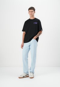 Black oversized T-shirt with a purple logo, light blue denim pants, and white sneakers with brown soles. Plain background.