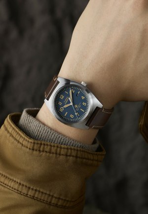 Randmikrofon sinise sihverplaadi, tan-numbrite, hõbedase korpuse ja pruuni nahast rihmaga, kantud randmel beeži sviitri ja pruuni jakkvarruka kohal.