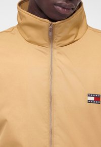 Крупен план на човек, облечен в бежово яке с цип и висока яка, с лого Tommy Jeans на гърдите.