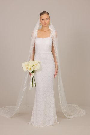 Donkerharige bruid in strapless kanten trouwjurk en lange geborduurde sluier, houdend een boeket van witte calla lelies, staand tegen een beige achtergrond.