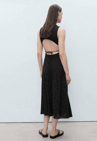 Robe noire avec un dos nu et un nœud ajustable, présentant un motif œillet, une jupe à mi-mollet et un corsage ajusté. Portée avec des sandales noires.