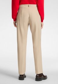 Pantalones beige a medida con una textura suave, presentan un frente plano y pliegues en la parte trasera, combinados con zapatos oscuros de suela gruesa.