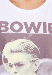 Valkoinen puuvillapaita, jossa on mustavalkoinen graafinen painatus kevyet hiukset omaavasta miehestä, joka wearing aurinkolasit; ylhäällä rohkeat "BOWIE"-tekstit.