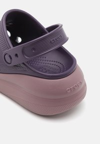 Des chaussures Crocs violettes avec un design texturé, présentant une semelle rose contrastante. Comprend une large sangle avec le logo de la marque et une semelle intérieure rembourrée.