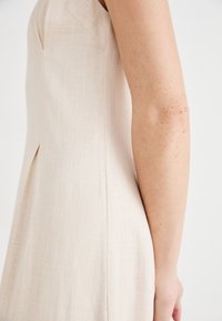 Robe sans manches beige en tissu texturé comme du lin, présentant un pli latéral et une silhouette ajustée, vue de côté.