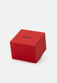 Raudona kartoninė dėžė su matinio paviršiaus tekstūra, kvadratinė forma, o viršuje išspausdinta žodžiu "HUGO" paryškintais juodais rašmenimis.