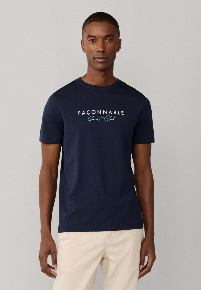 Man draagt een marineblauw "Façonnable Yacht Club" T-shirt en lichtbeige broek, staand tegen een effen lichtgrijze achtergrond.