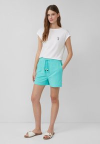 T-shirt bianca a maniche arrotolate con una piccola grafica sul petto, abbinata a pantaloni corti turchesi con cordoncino e dettagli di perline. I sandali sono bianchi.