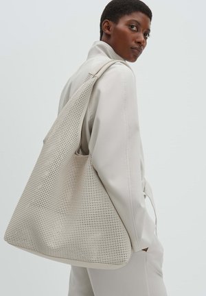 Schwarze Frau mit beigem Trenchcoat trägt eine große beige perforierte Einkaufstasche über der Schulter und blickt über ihre linke Schulter.