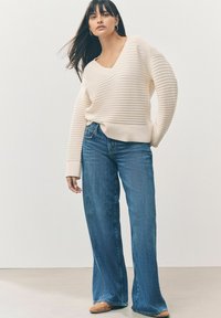 Maglione a coste crema con scollatura a V, abbinato a jeans blu a gamba larga. Il modello indossa scarpe slip-on beige, su uno sfondo neutro.