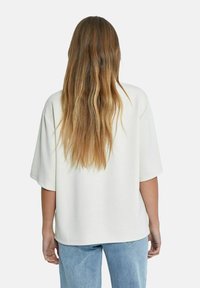 Suéter de corte amplio en color crema claro, con un ajuste relajado y mangas cortas anchas. En la parte de atrás tiene una superficie suave y texturizada. Combinado con unos jeans de color azul claro.