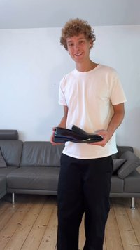 Crna kožna cipelica bez kopčanja s sjajnim finishem, izduženog oblika i minimalističkog dizajna, drži osoba u bijeloj košulji.