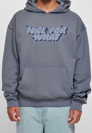 Person trägt grauen Kapuzenpullover mit dem Schriftzug „NICE FOR WHAT“ in stilisierter Schrift auf der Vorderseite und einer kleinen Grafik am linken Ärmelsaum.