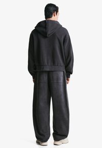 Eine Person trägt einen lockeren, dunkelgrauen Kapuzenpullover und passende weite Hosen. Sie steht mit dem Rücken zur Kamera, weiße Sneakers sind sichtbar.