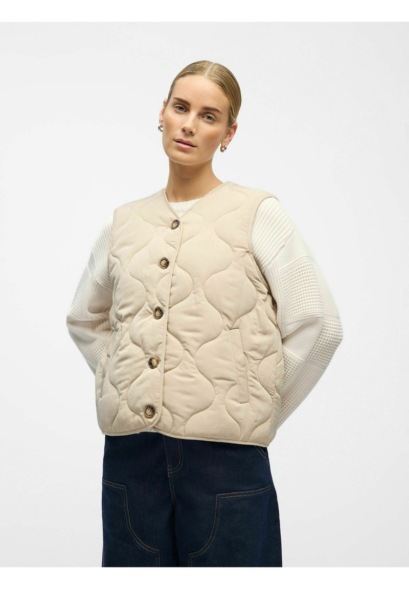 Beige gewatteerde vest met een ronde halslijn, voorzien van vijf knoopsluitingen, zijzakken en een zachte textuur. Draagt over een wit gebreid shirt.