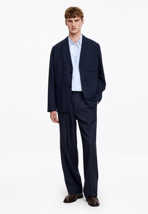 Homme portant un costume décontracté bleu marine foncé avec une chemise bleu clair boutonnée et des chaussures marron foncé, debout avec une main dans la poche sur fond blanc.