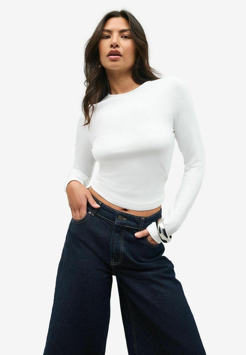Witte blouse met lange mouwen, gecombineerd met donkerblauwe high-waisted denim wijde pijpenbroek. Een zilveren armband siert de pols en voegt een metalen accent toe.