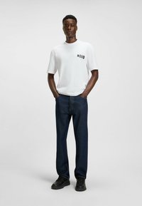 Young man standing with hands in pockets wearing white Hugo t-shirt, dark blue jeans, and black sneakers on plain gray background.