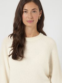 Kremowy sweter o miękkiej fakturze i rękawach raglanowych. Modelka nosi złote kolczyki w kształcie kół. Prosty krój z okrągłym dekoltem.